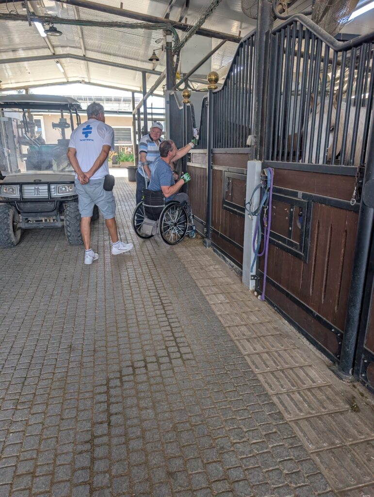 מבט לאורך מעבר רחב ומרוצף באורווה מקורה. אדם בכיסא גלגלים ואדם מבוגר המסתייע בקביים נמצאים ליד תאי הסוסים, לצד סוקר נגישות הלובש חולצה לבנה עם הלוגו של "נגישות ישראל". משמאל חונה רכב גולף קטן.