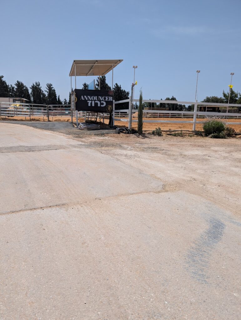 רחבה חיצונית של חוות סוסים עם דרך עפר סלולה ומפולסת בחזית. ברקע נראית גדר עץ לבנה המקיפה את זירת הרכיבה, ומבנה מוגבה ומקורה עם שלט שחור שעליו כתוב "ANNOUNCER כרוז".