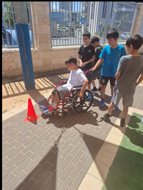 ארבעה ילדים עומדים סביב ילד חמישי היושב בכיסא גלגלים אדום על שביל מרוצף. הילד בכיסא הגלגלים לובש חולצה לבנה ומכנסיים קצרים בדוגמה שחור-לבן. הילדים שעומדים סביבו נראים בגילו. אחד מהם עומד מאחורי כיסא הגלגלים. מול כיסא הגלגלים על השביל ניצב קונוס כתום. ברקע נראית גדר ברזל, עצים ומבנים עירוניים מודרניים. השמש מאירה והצללים ברורים.
