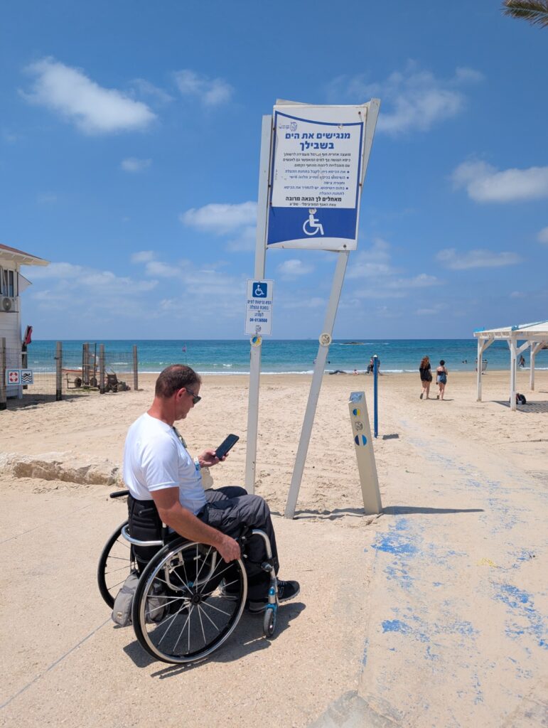 אדם המשתמש בכיסא גלגלים נמצא בכניסה לחוף נווה ים, סמוך לשלט המציין את שירותי הנגישות בחוף. מהשביל הנגיש מובילה דרך אל קו המים, וברקע נראים הים, החוף החולי, מתרחצים ושמיים כחולים עם עננים.