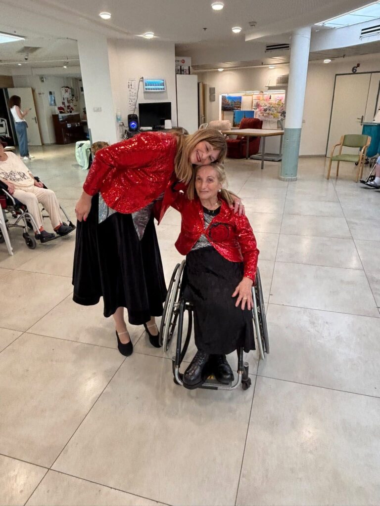 שתי נשים מחייכות ומצטלמות יחד באירוע ברמת יוחנן, אחת מהן יושבת בכיסא גלגלים ושתיהן לבושות בבגדים חגיגיים. 
