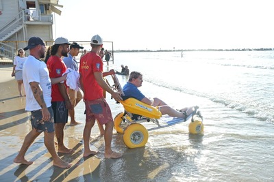 A man sits in an amphibious beach wheelchair as several people help him enter the sea. The wheelchair is equipped with large wheels and a yellow flotation support. A lifeguard tower and the shoreline are visible in the background.