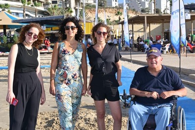 Four people pose together at a beach, including a man seated in a wheelchair. Three women stand beside him, and one holds a red phone. Beach umbrellas, flags, and the sea are visible in the background.