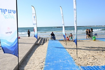 A blue accessible beach mat leads from the shore toward the sea, with a person in a beach wheelchair near the water. Access Israel flags stand along the path, and beachgoers are visible in the background.
