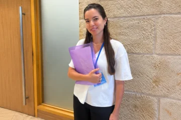 אישה עומדת במסדרון בכנסת, ליד דלת עץ וקיר אבן, לבושה בחולצה לבנה ומכנסיים שחורים, מחזיקה תיקייה סגולה ועונדת תג זיהוי כחול.