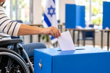 אדם היושב בכיסא גלגלים ומכניס פתק הצבעה לקלפי כחולה, על רקע דגל ישראל ועמדות הצבעה.