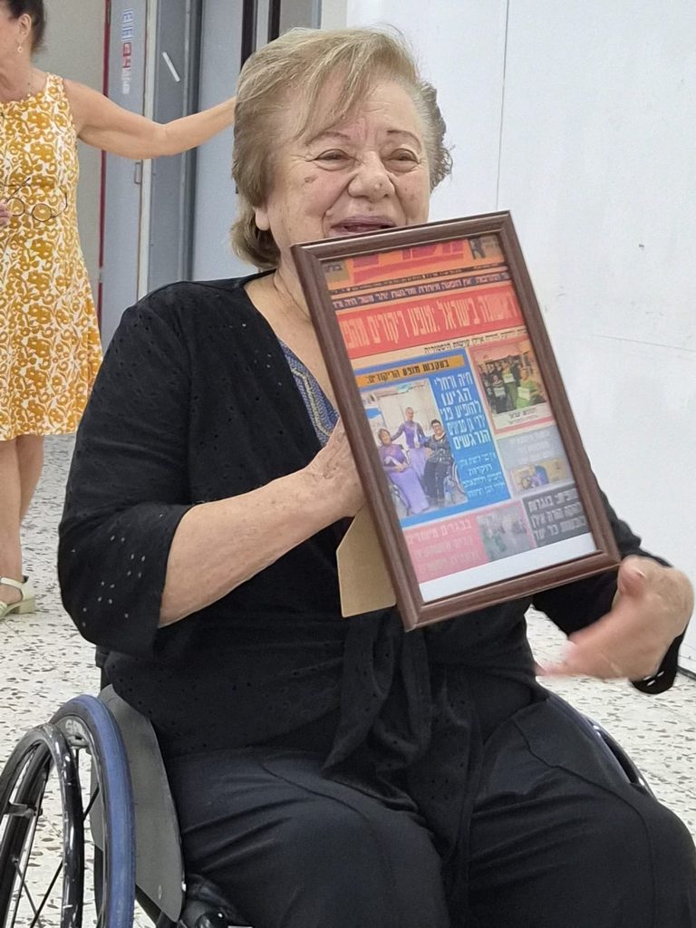 אישה בכיסא גלגלים מציגה מסגרת ובה גזירי עיתונות ותמונות אישיות במסגרת סדנת „סיפורו של חפץ” למתנדבי נגישות ישראל צפון.