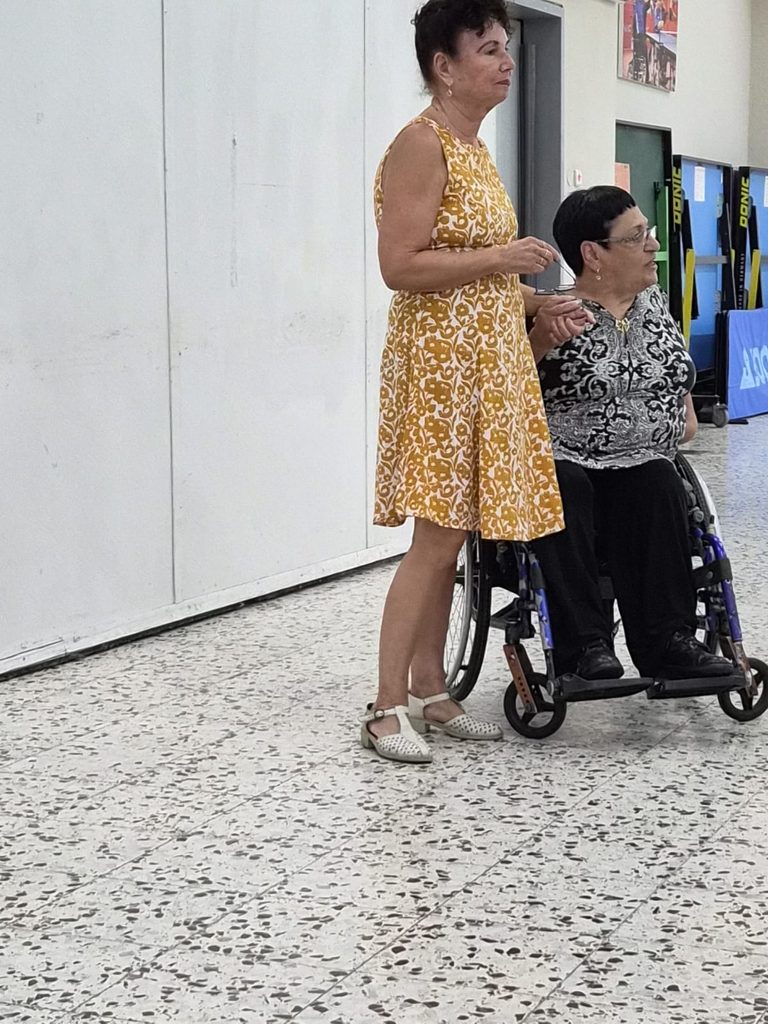 מתנדבת בשמלה צהובה מגישה שי לאישה בכיסא גלגלים במהלך מפגש מתנדבי נגישות ישראל צפון.
